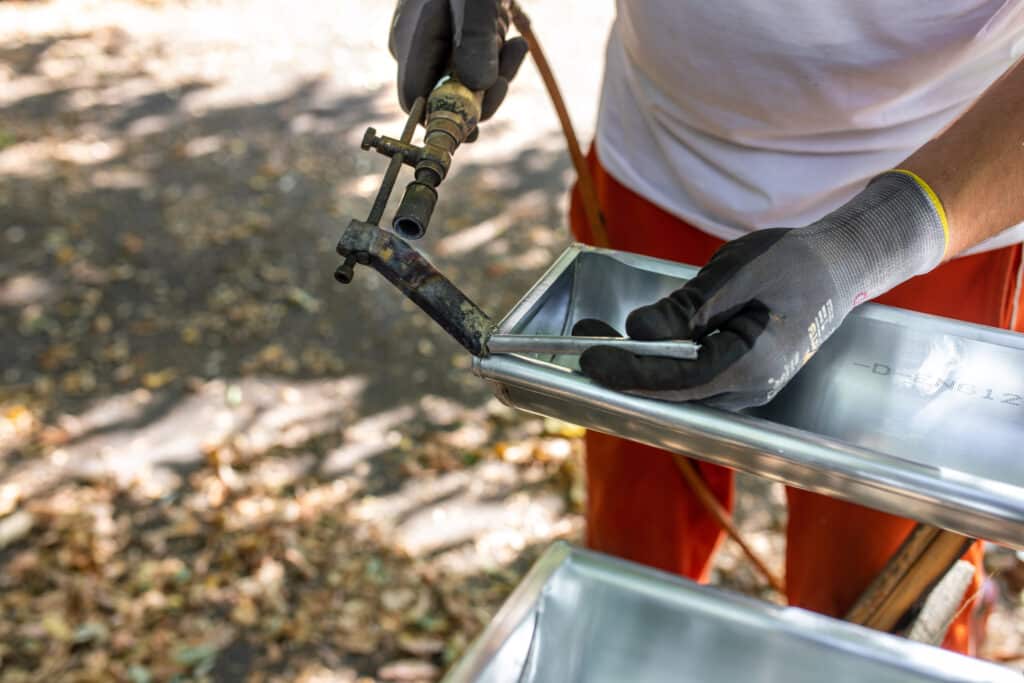 Main gantée tenant un chalumeau et insérant une tige métallique dans un bac en acier inox marqué "-D-ENG12". Bricolage extérieur.