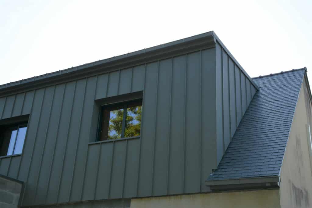 Façade moderne en bardage métallique anthracite avec fenêtre reflétant des arbres, et toiture en ardoise sombre.