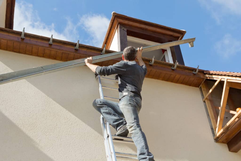 Homme sur échelle installant une longue gouttière métallique sur la façade d'une maison avec boiseries sous un ciel bleu.