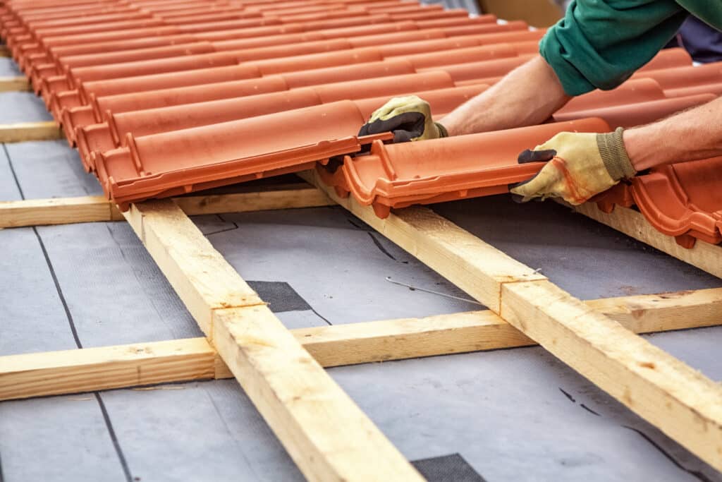 Mains gantées posant des tuiles de toit en terre cuite sur des lattes en bois et une membrane étanche. Construction d'une toiture.
