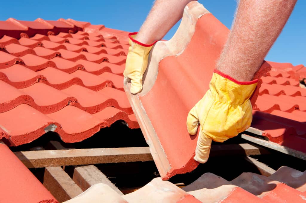Mains gantées d'un couvreur posant une tuile rouge sur un toit en construction sous un ciel bleu clair.