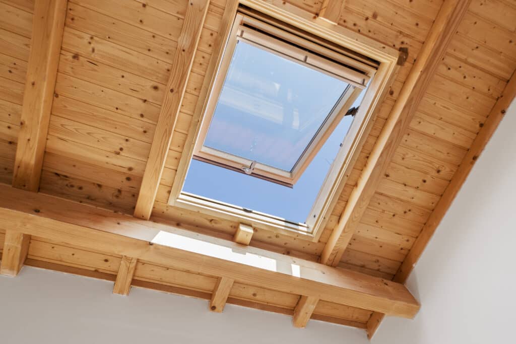 Plafond mansardé en bois clair avec poutres, sous un puits de lumière Velux ouvert. Ciel bleu visible par la lucarne, intérieur lumineux.