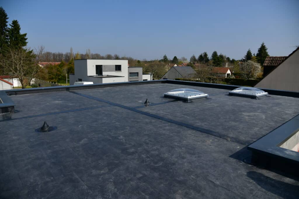 Toit plat en bitume noir avec deux lucarnes et des empreintes de pas. Arrière-plan de maisons modernes et traditionnelles sous un ciel bleu dégagé.