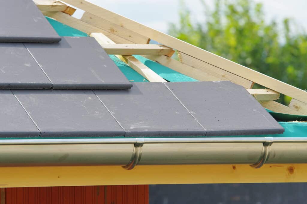 Toiture en construction avec tuiles grises, membrane verte, charpente en bois et gouttière métallique. Ciel clair et arbres en arrière-plan.