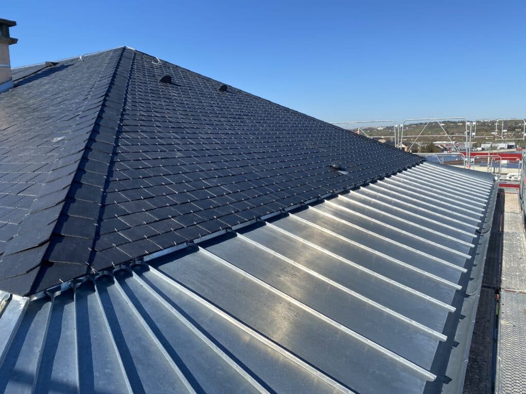 Couverture Ardoise et Zinc: L'art du toit à joint debout Couverture en ardoise noire neuve, fixée par crochets, raccordée à un toit en zinc à joint debout. Scaffolding visible sous ciel bleu éclatant.