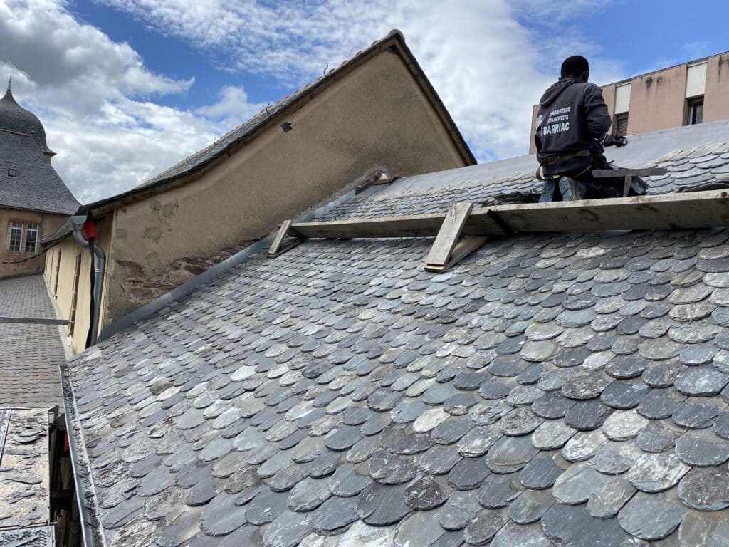 Couvreur Barriac : Réparation de toit en ardoise traditionnelle Artisan COUVERTURE ÉTANCHÉITÉ BARRIAC travaillant sur un toit ancien en ardoise grise (écailles). Détail de la pose.