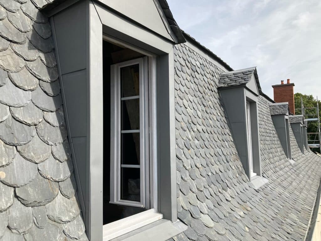 Toiture ardoise écaillée : pose de lucarnes et zinc Longue toiture mansardée en ardoise écaille. Quatre lucarnes à joues de zinc gris. La première montre un châssis blanc ouvert.